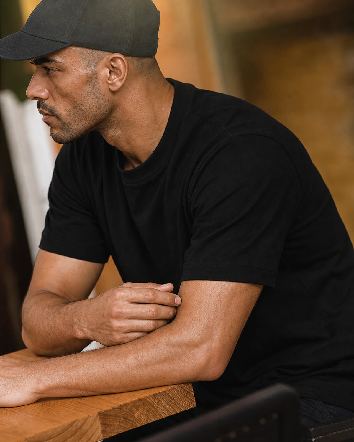 Man in the black heavyweight cotton tee and cap, leaning on a table
