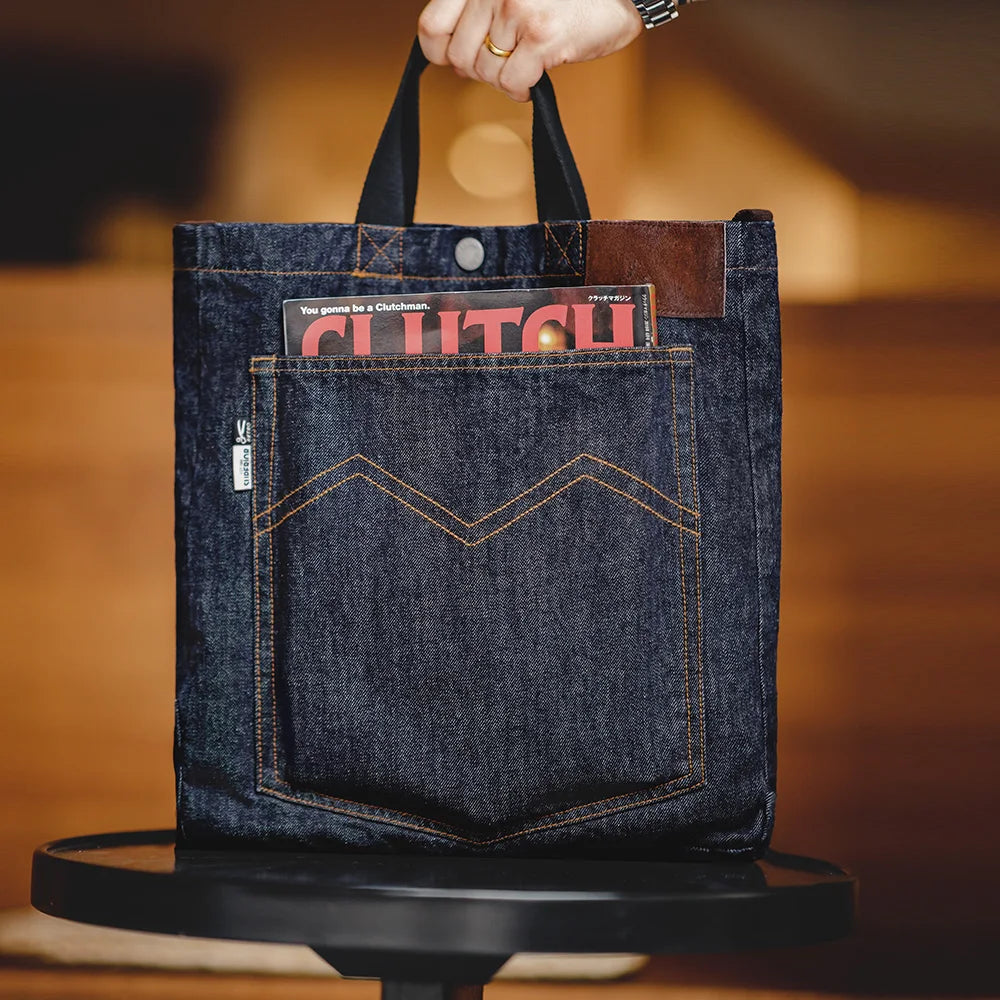 Navy denim tote bag carried by its black top handles, brown leather patch at the upper corner